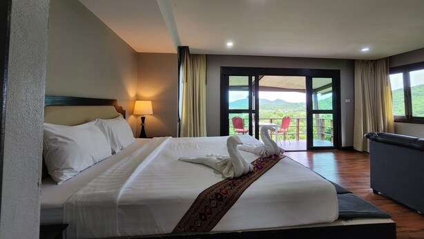 Een hotelkamer met een groot bed, een nachtkastje met een lamp, uitzicht op de natuur door een schuifdeur, en een balkon met rode stoelen.