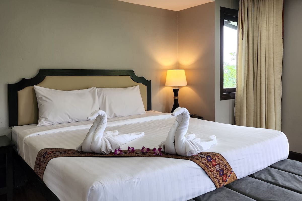 A hotel room with a bed, a nightstand, a lamp, a window with curtains, and a view of greenery outside.