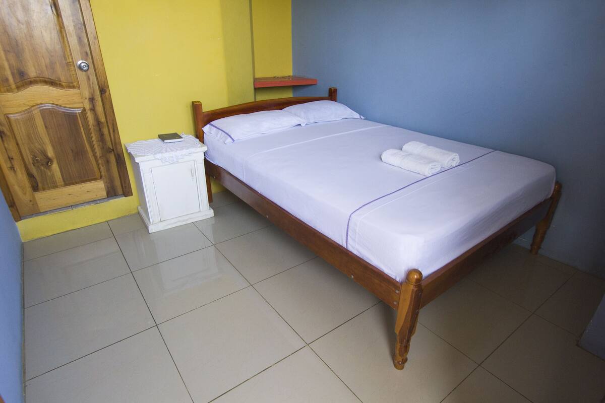 A neatly made bed with white linens and two rolled towels, a wooden nightstand with a lamp, and a wooden door.