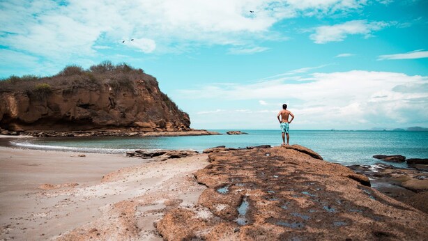 A person standing on a rocky outcrop overlooking a beach with clear skies and calm waters. - Hostal Alcazaba (Puerto Lopez)