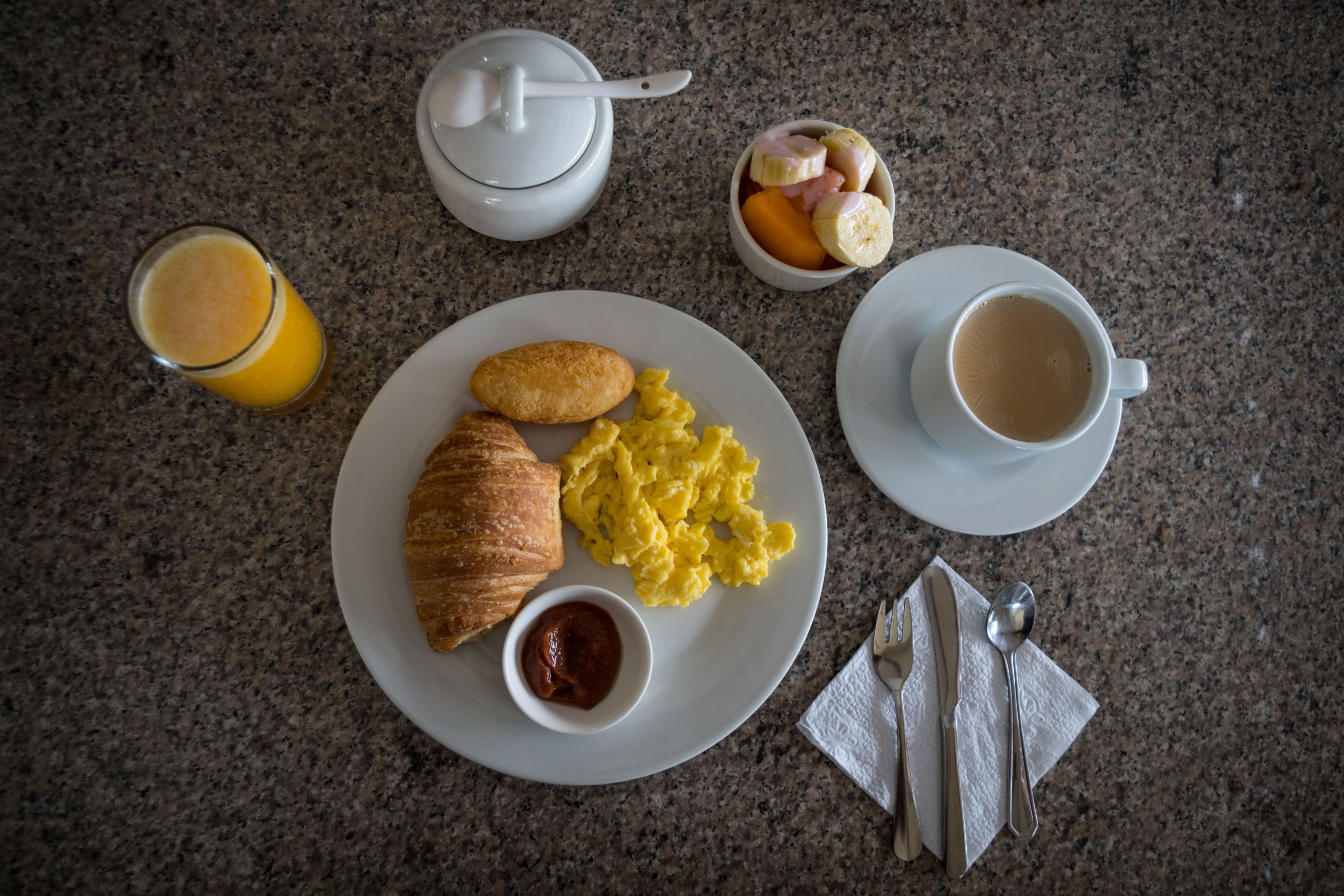 A breakfast setting with a croissant, scrambled eggs, a glass of orange juice, a cup of coffee, and a bowl of fruit on a granite countertop.