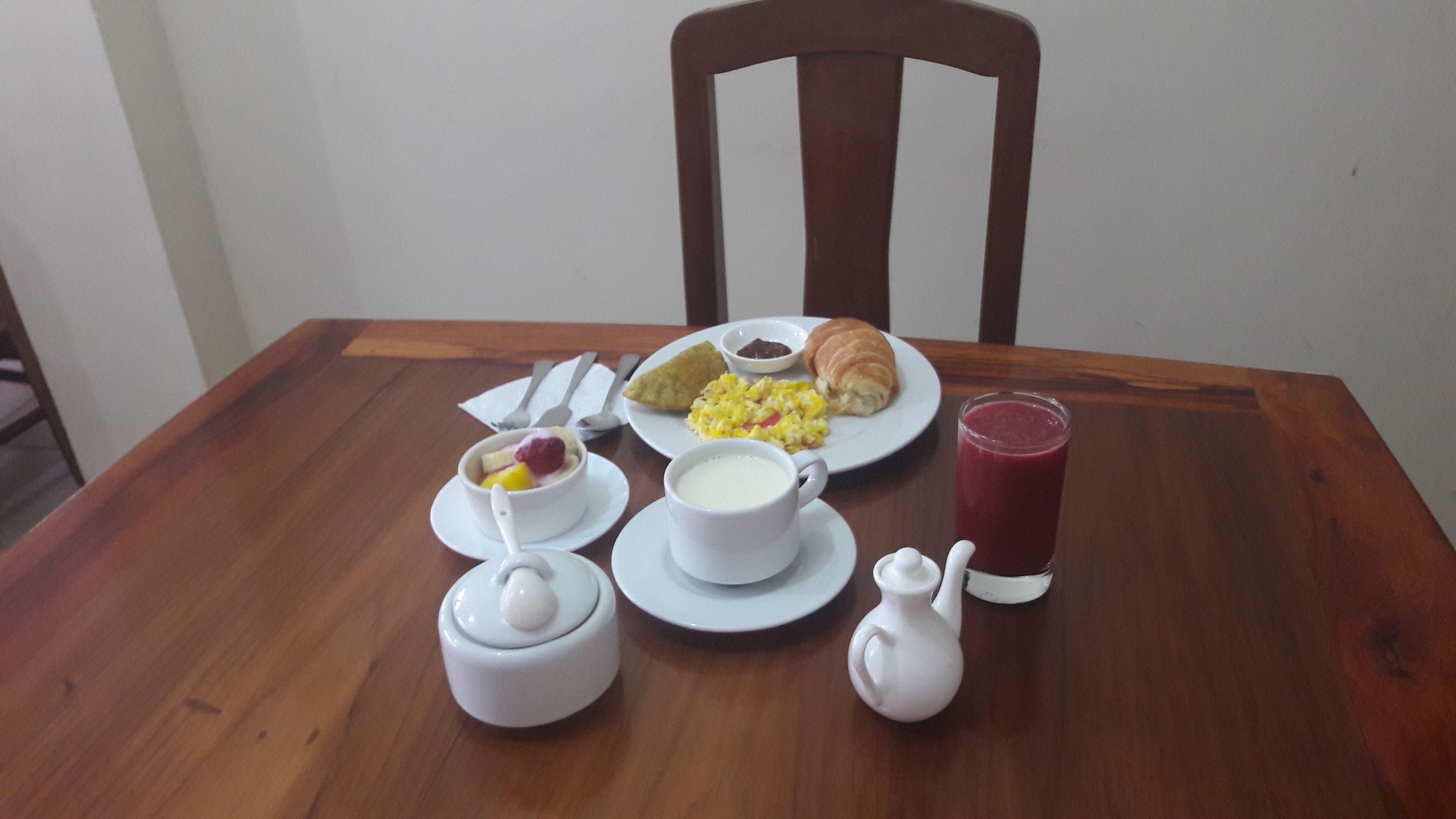 Una mesa de desayuno con un croissant, huevos revueltos, un bol de fruta, un vaso de jugo y una taza de leche.