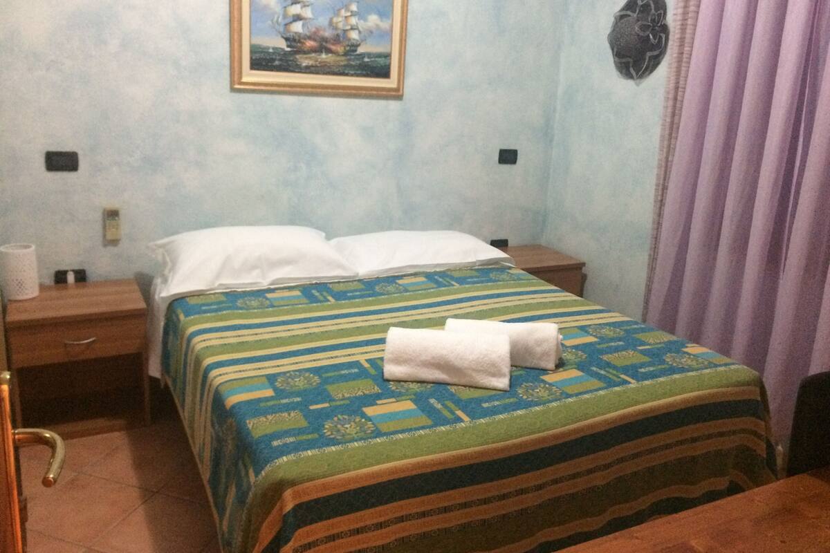 A hotel room with a bed, a nightstand, a lamp, a painting on the wall, and a curtain.