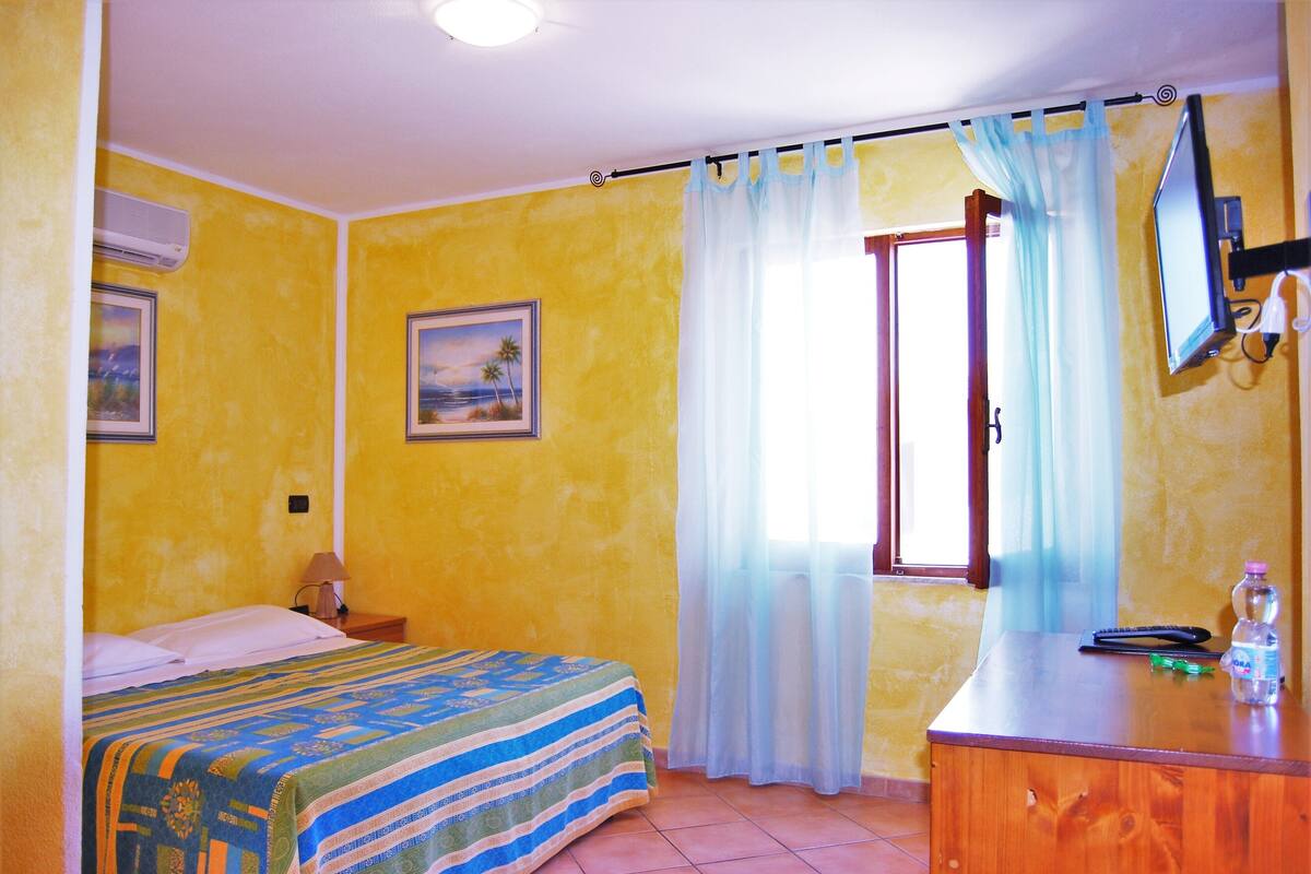 A room with a bed, a wooden dresser, a desk, a lamp, a picture on the wall, and a window with curtains.