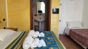 Habitación de hotel con cama, mesita de noche, baño con lavamanos y espejo, y una lámpara de pared.