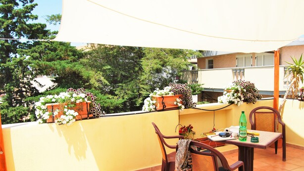 Un balcón con una mesa y sillas, plantas en macetas y vistas a árboles y edificios.