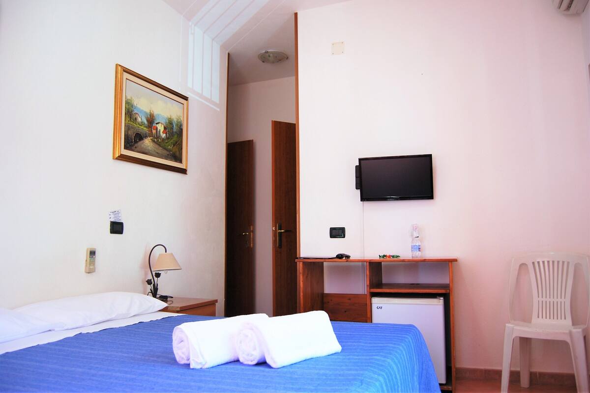 A hotel room with a bed, a desk, a television, a chair, and a painting on the wall.