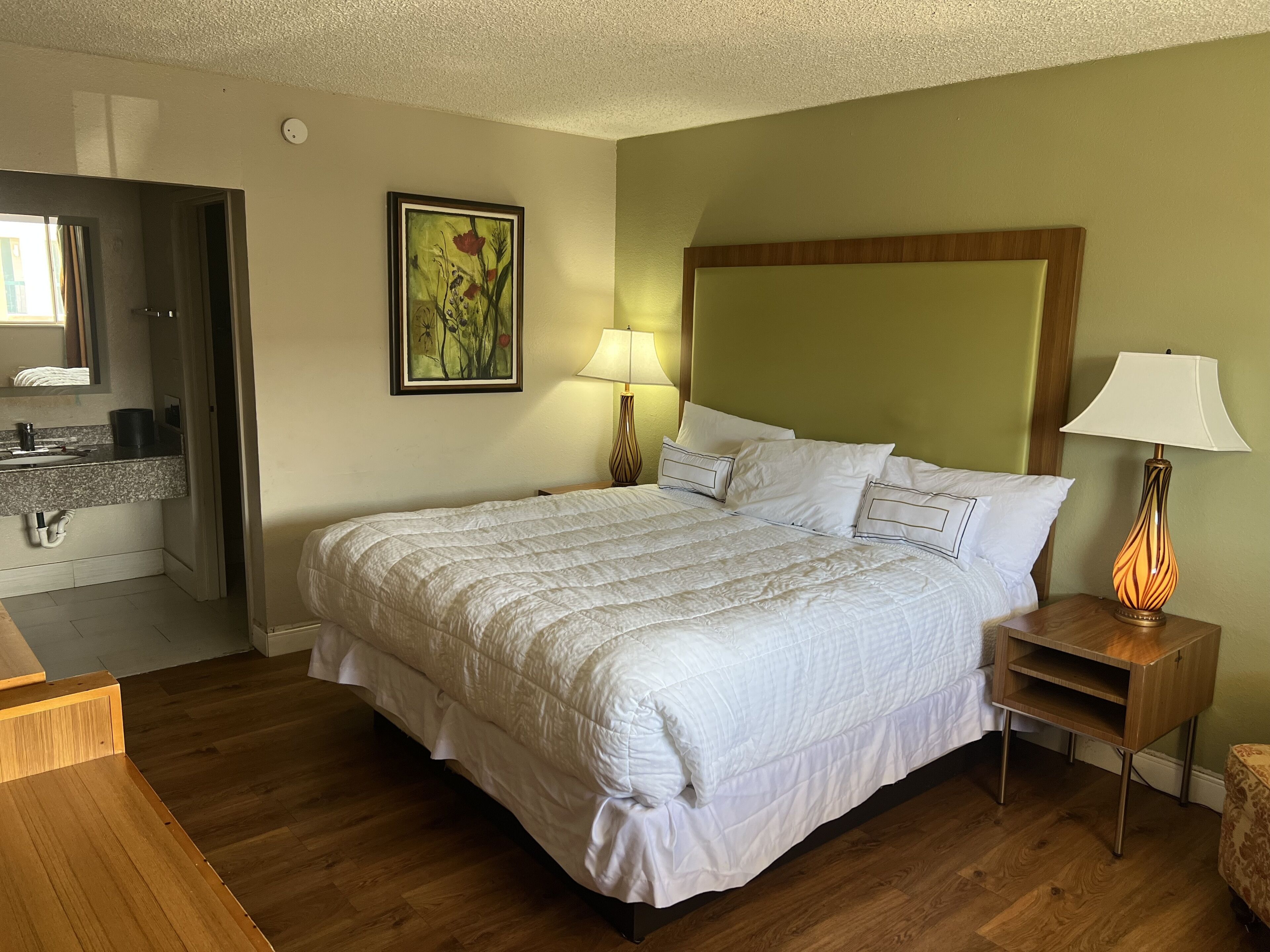 Een hotelkamer met een groot bed, een houten nachtkastje, een bedlampje, een badkamer met een wastafel en spiegel, en een schilderij aan de muur.