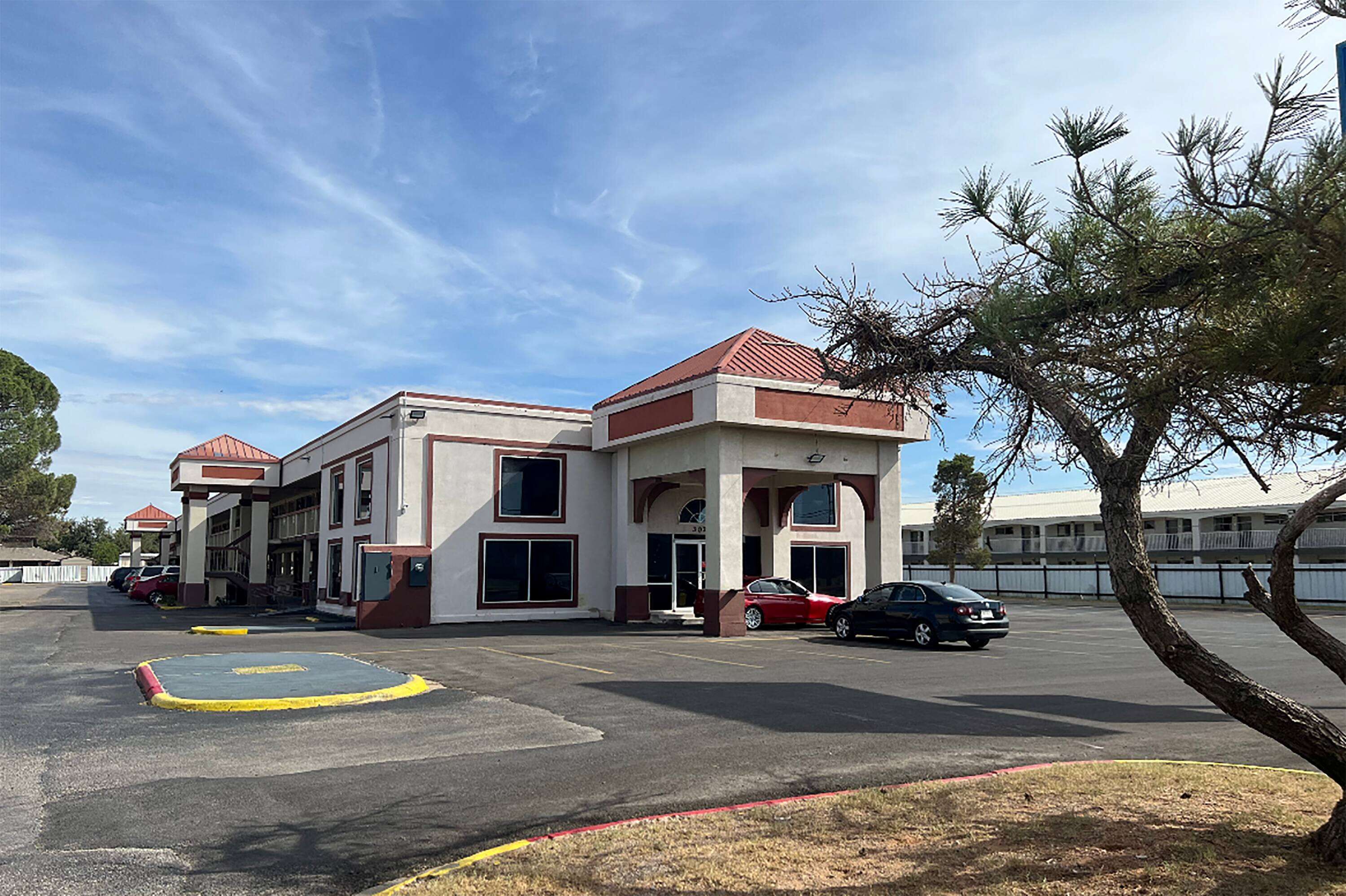 A two-story building with a red roof and large windows. There are cars parked in front and a tree to the right.