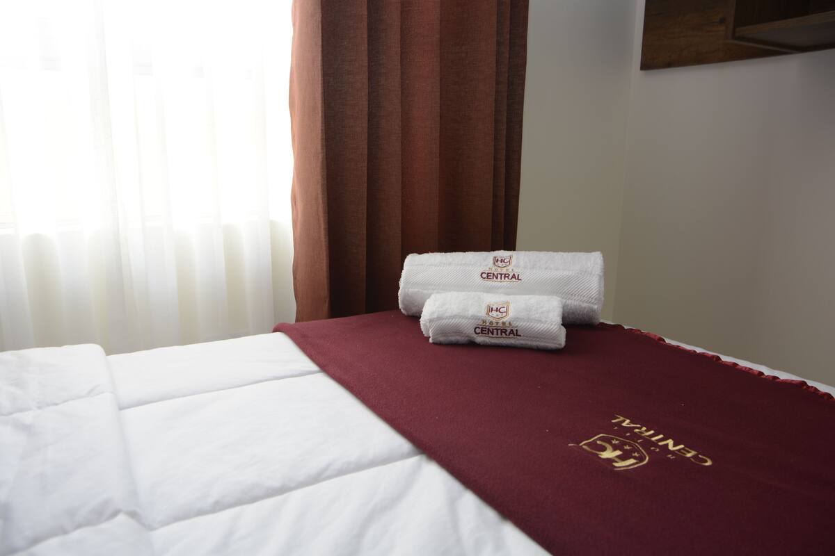 A hotel room with a neatly made bed, a red blanket with the hotel logo, and a window with curtains.