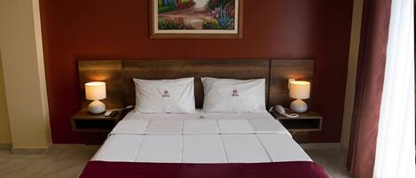 Una habitación de hotel con una cama grande, dos mesitas de noche con lámparas, un cuadro en la pared y una ventana con cortinas.