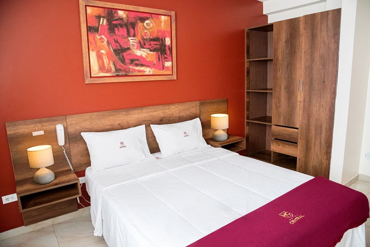 A hotel room with a large bed, two bedside lamps, a wooden headboard, a red accent wall, and a large wardrobe.