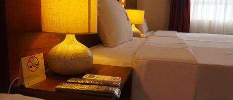 A hotel room with a bed, a nightstand with a lamp, a remote control, and a no-smoking sign.