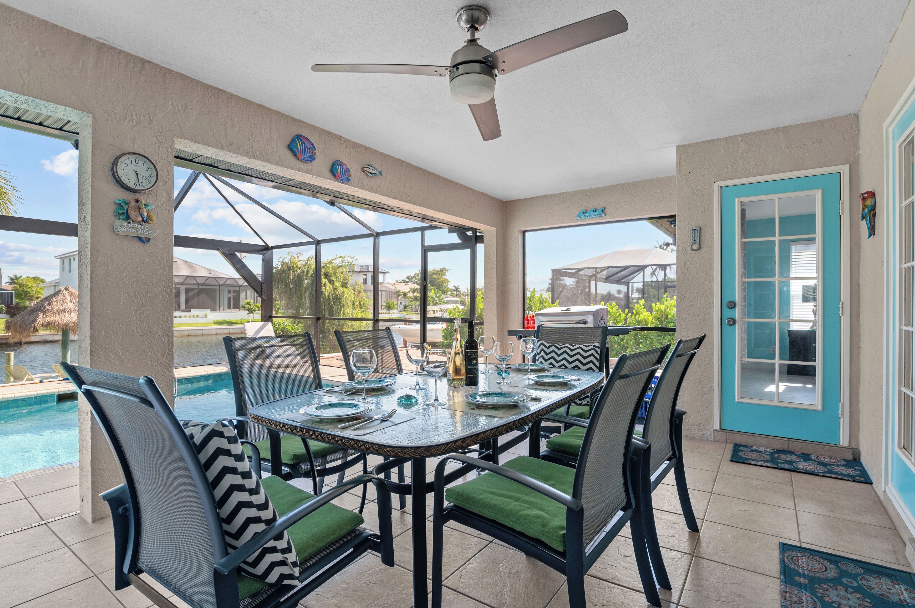 Un coin repas avec une table dressée pour quatre personnes, des chaises avec des coussins verts, un ventilateur de plafond et une vue sur une piscine et des maisons.