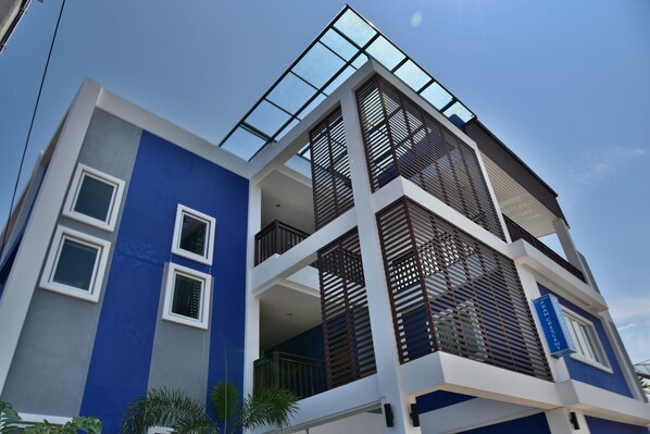 A modern multi-story building with a blue facade, large windows, and a balcony with wooden slats.