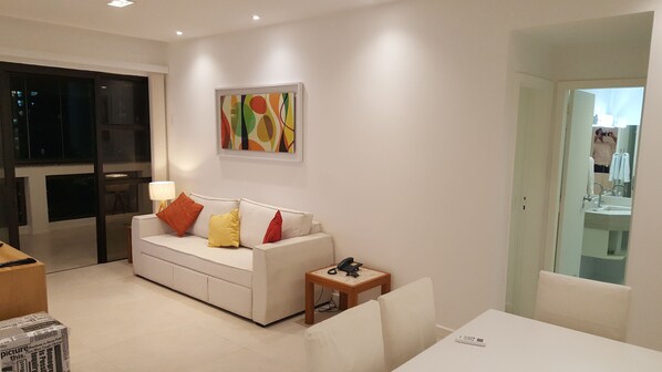 Un salon moderne avec un canapé blanc, des coussins colorés, une petite table avec un téléphone et un tableau accroché au mur.