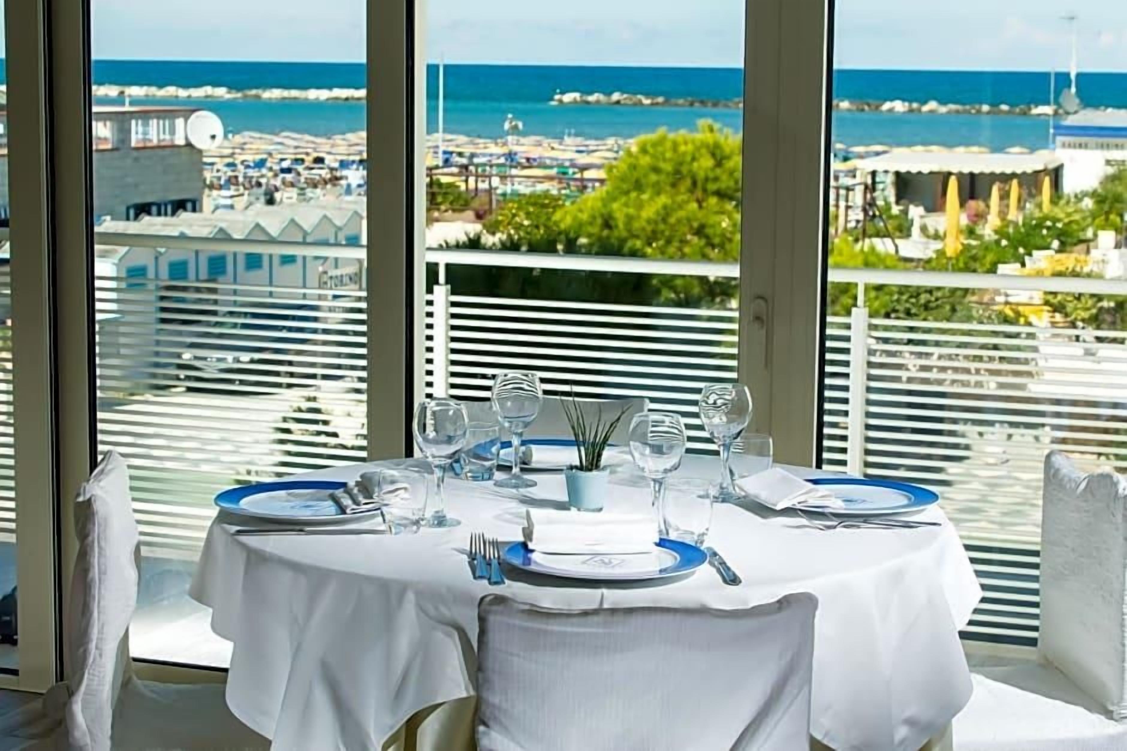 Une table à manger dressée avec des assiettes, des couverts et des verres, offrant une vue sur la mer et les bâtiments en arrière-plan.