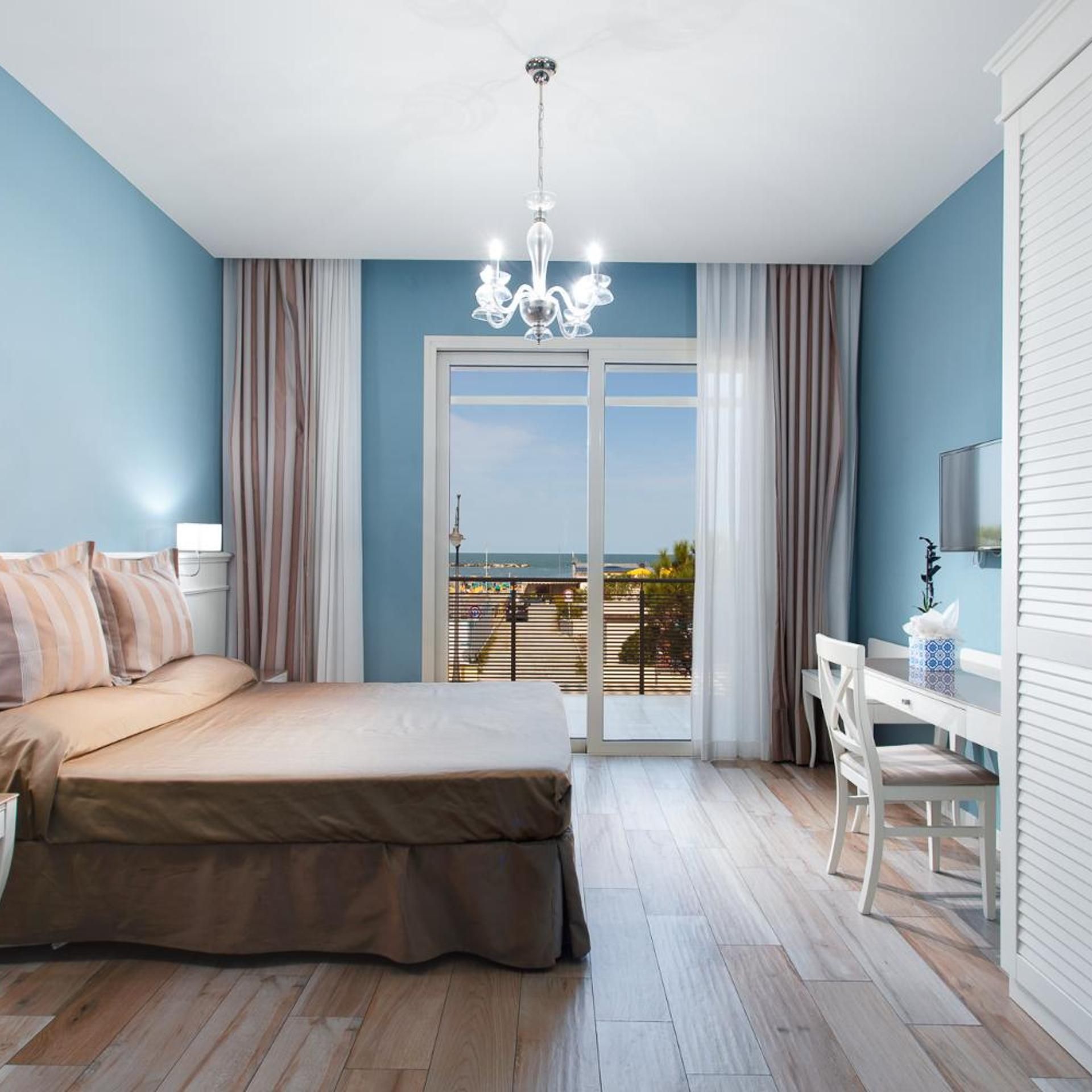 A bedroom with a bed, a desk, and a view of the sea through a glass door.