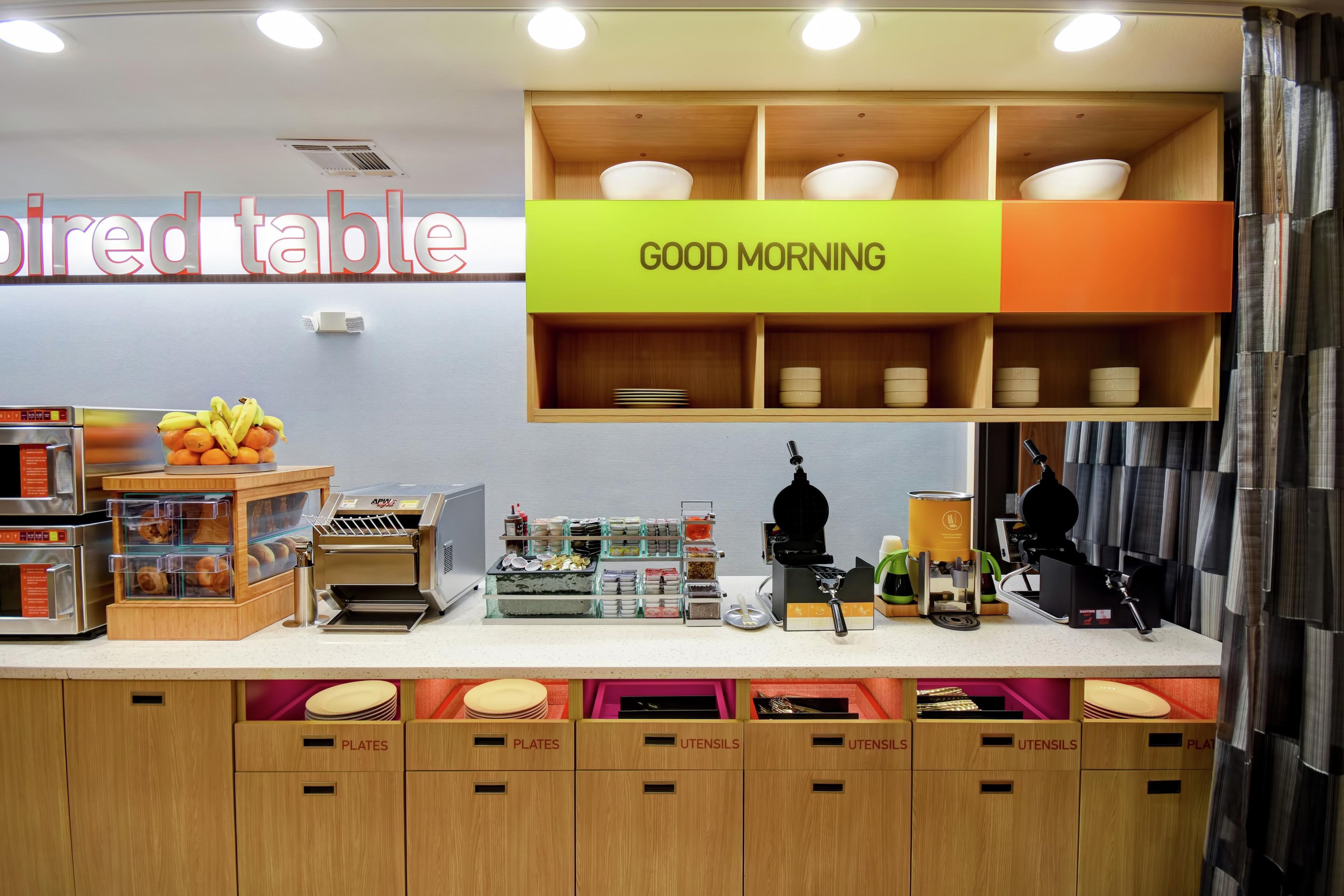 A breakfast area with a coffee machine, a fruit display, and labeled storage for plates and utensils.