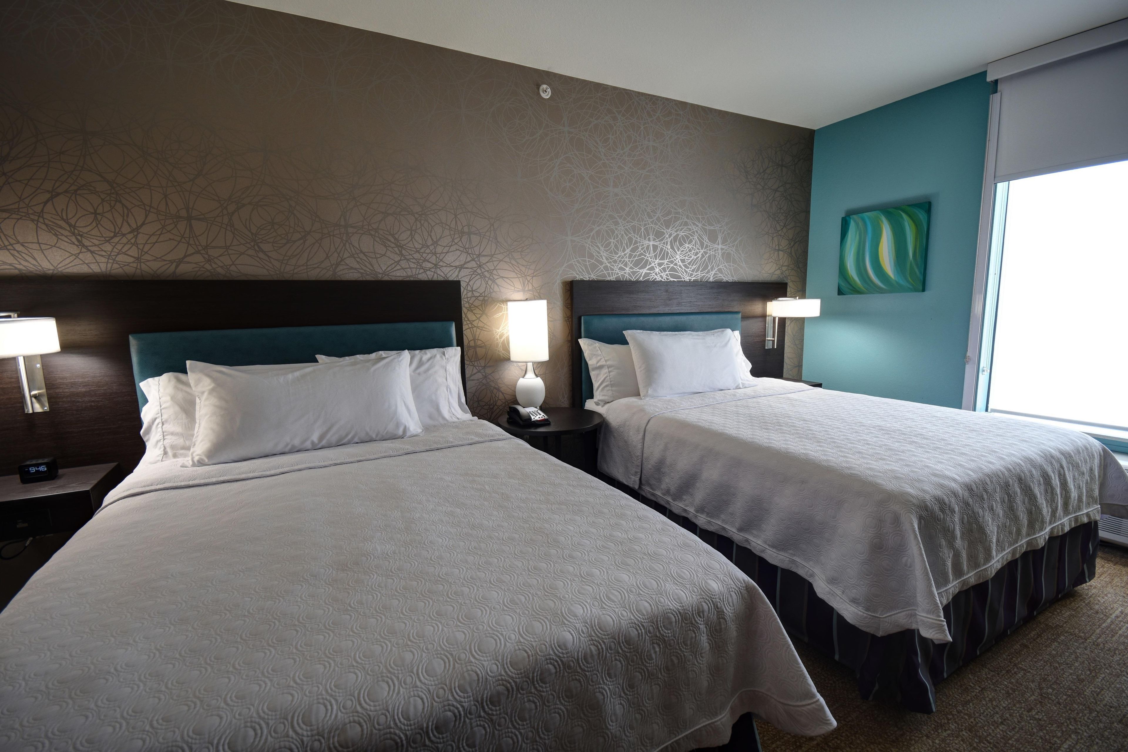 A hotel room with two beds, each with a headboard, bedside lamps, and a remote control on the nightstand.