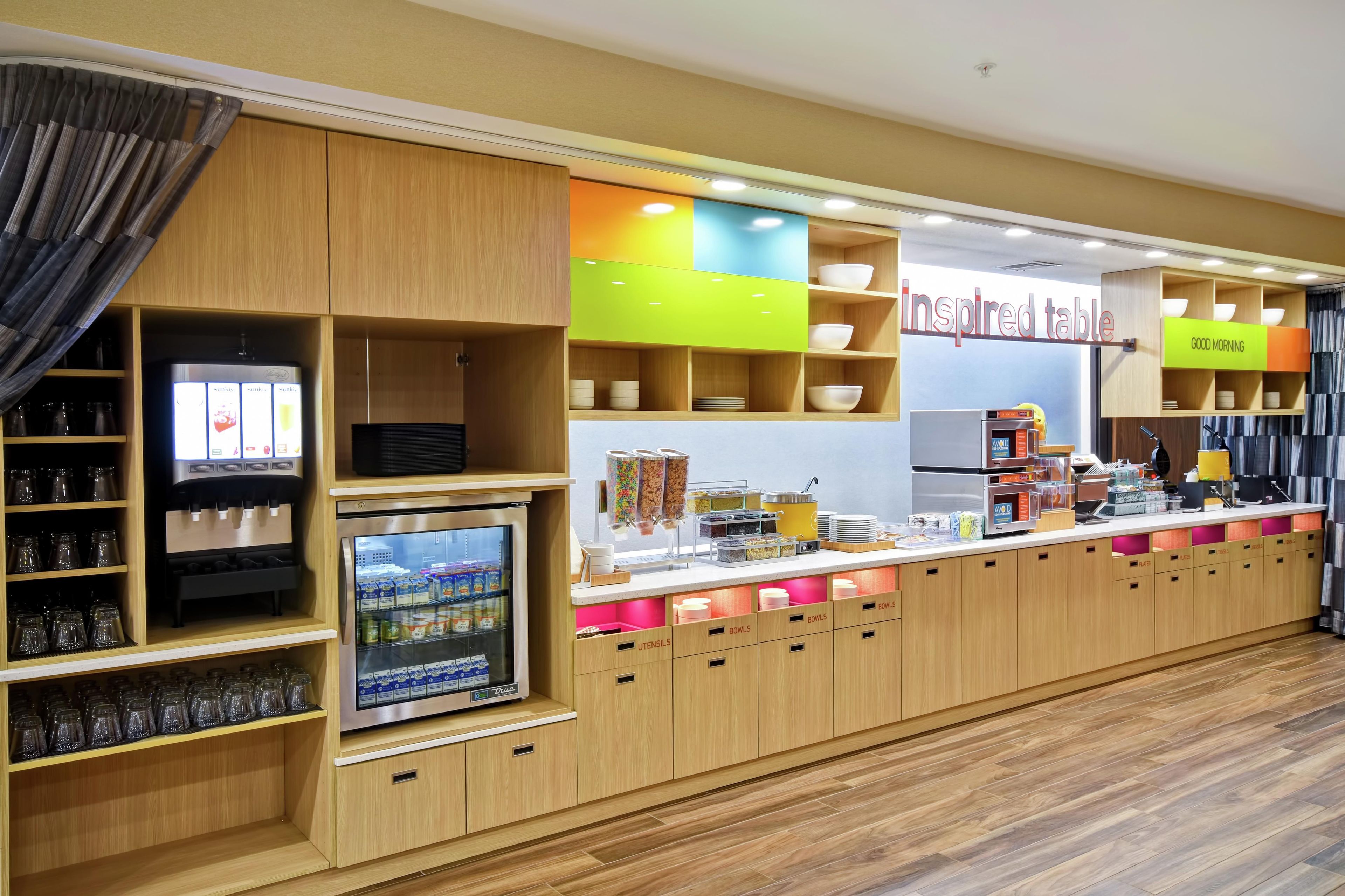 A self-service breakfast area with a coffee machine, refrigerated drinks, and various food containers.