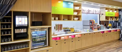 A self-service breakfast area with a coffee machine, refrigerated drinks, and various food containers.