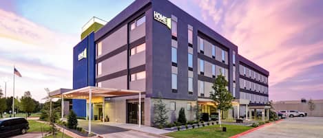 A modern hotel building with a 'HOME' sign, a covered entrance, and a parking area.