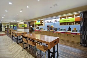 Un restaurant moderne avec des tables et des chaises en bois, un comptoir orné d’une pancarte « Bonjour » et une autre pancarte, « Table d’Inspiration », au-dessus du comptoir.