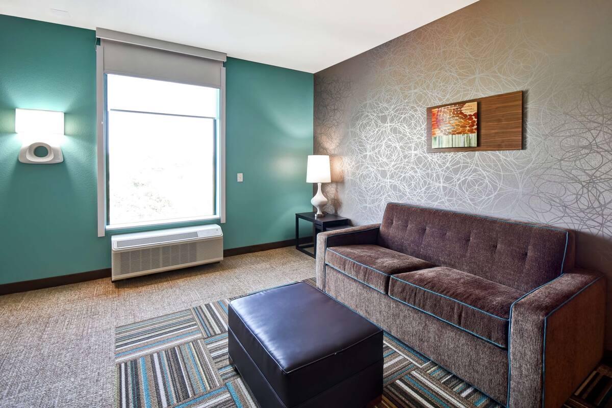 A modern living room with teal walls, a brown sofa, a black ottoman, and a patterned rug.