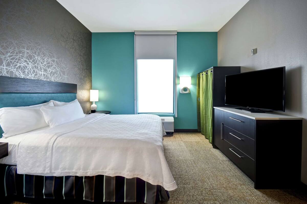 A hotel room with a bed, a TV, a dresser, a lamp, and a window.