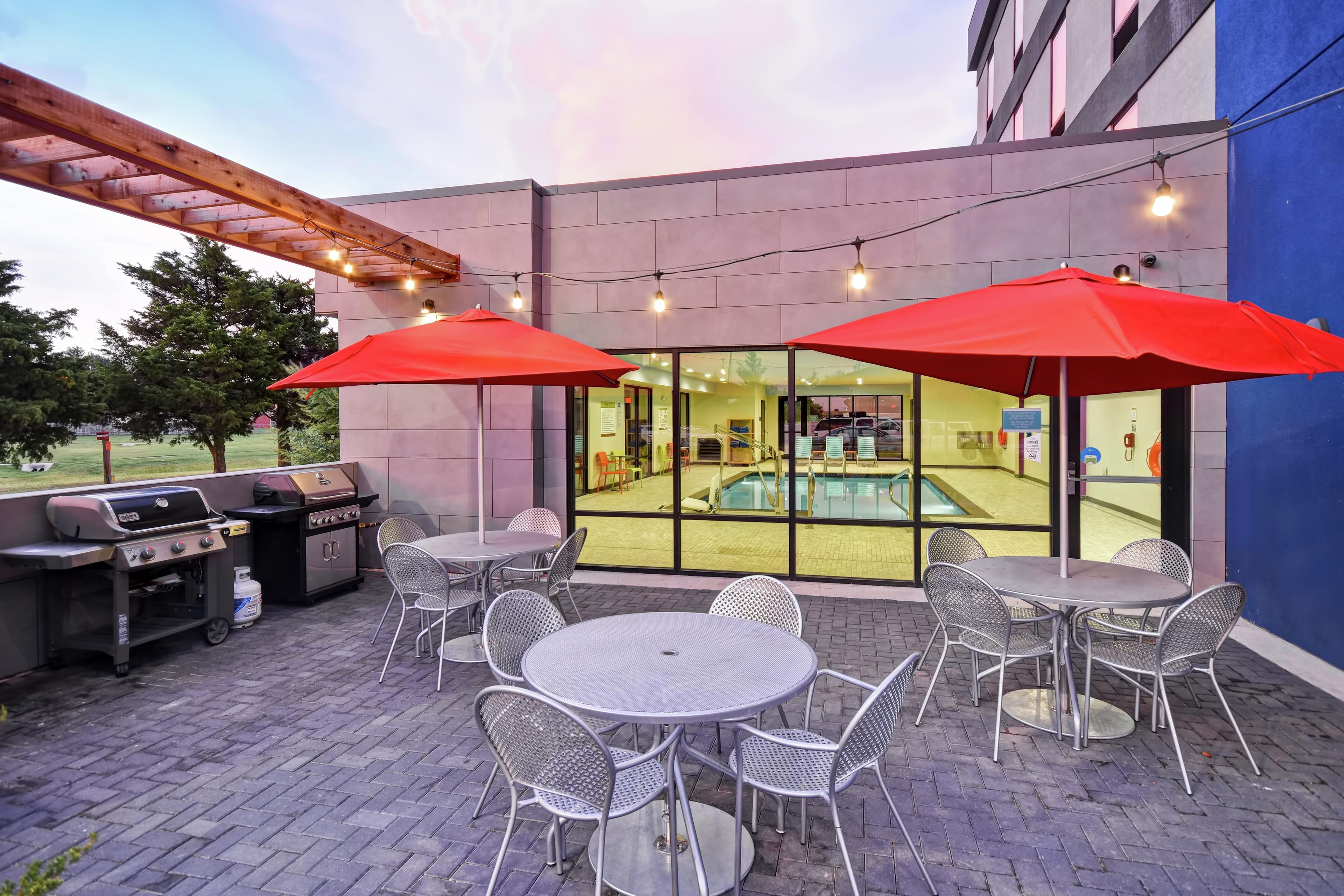 Un patio exterior con mesas y sillas de metal, una parrilla y una piscina visible a través de las puertas de vidrio.