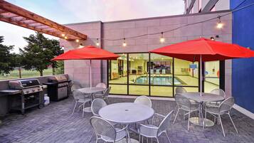 Un patio exterior con mesas y sillas de metal, una parrilla y una piscina visible a través de las puertas de vidrio.