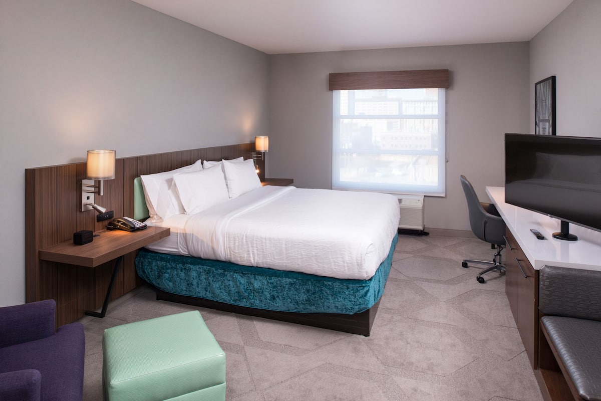 A hotel room with a bed, desk, chair, and a window with a view of buildings.