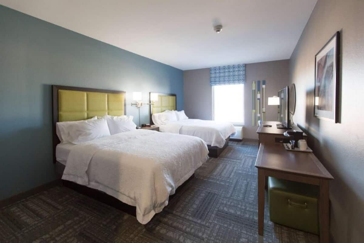 A hotel room with two beds, a desk, a chair, a small table, and a window with blinds.