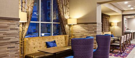 A hotel lobby with a seating area featuring a sofa and chairs, a dining area with tables and chairs, and a large window with curtains.