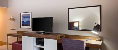 A modern hotel room with a wooden desk, a flat-screen TV, a mirror, a purple chair, and a red ottoman.