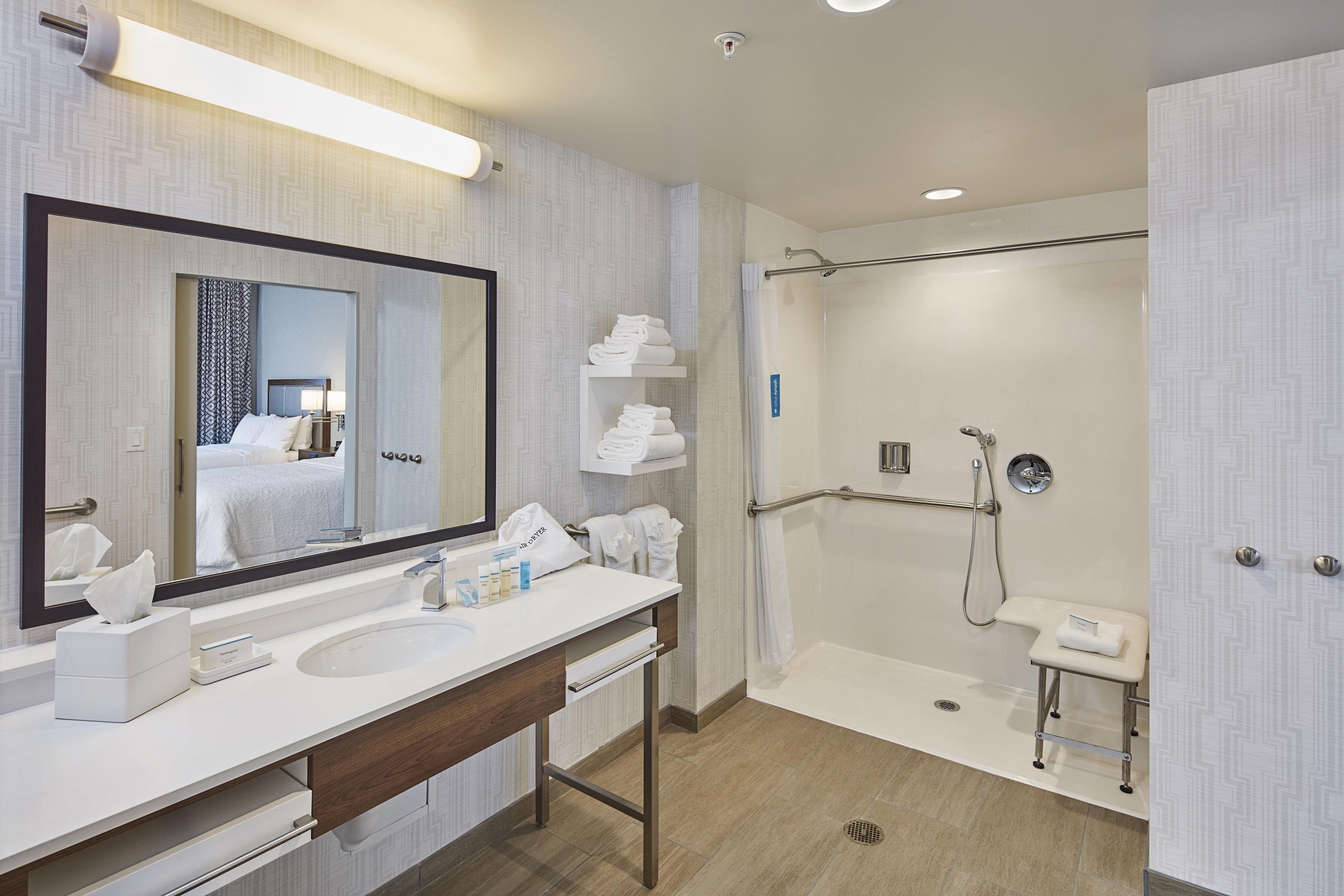 A modern bathroom with a large mirror, a sink, a shower with a grab bar, and neatly folded towels.