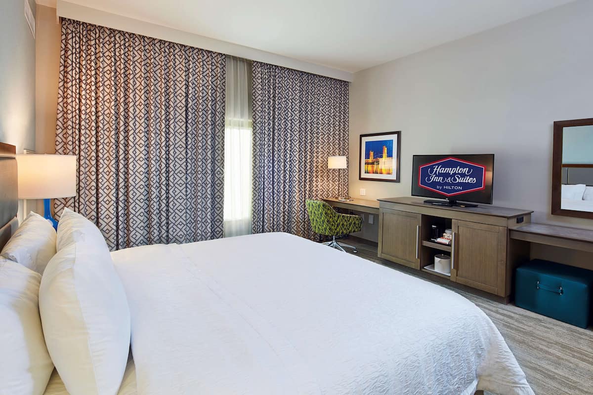 Hotellihuone, jossa on suuri sänky, työpöytä, jossa on televisio, kuviolliset verhot ja Hampton Inn & Suites -kyltti.