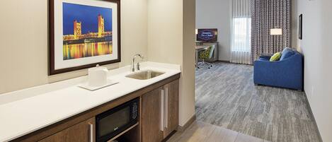 A hotel room with a kitchenette, a large window with curtains, a framed picture of a bridge, a flat-screen TV, and a blue sofa.
