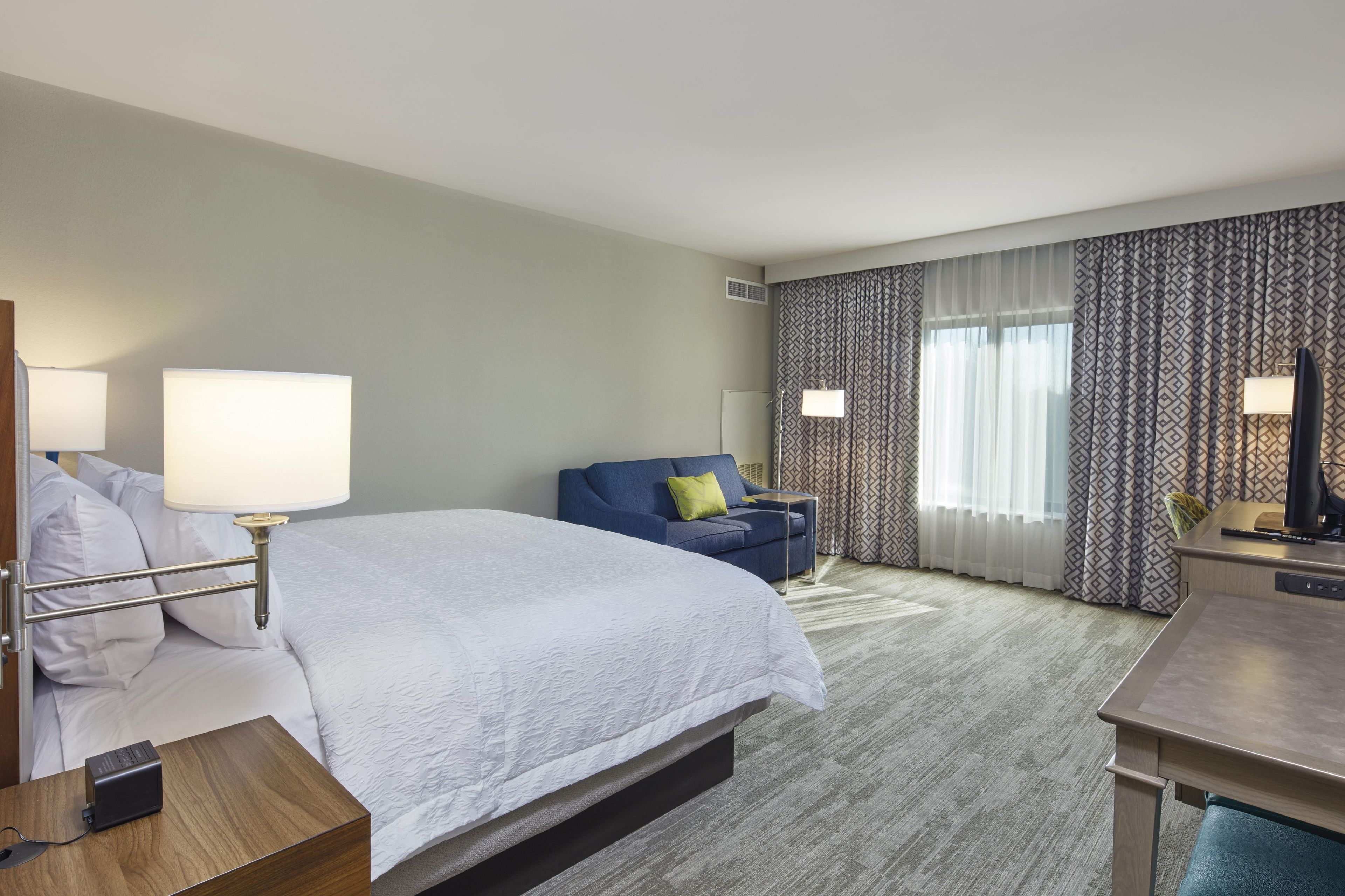 A hotel room with a bed, a desk, a chair, a lamp, and a window with curtains.