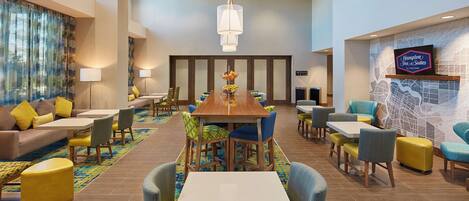 A spacious hotel lobby with a central dining area, comfortable seating, and a television mounted on the wall.