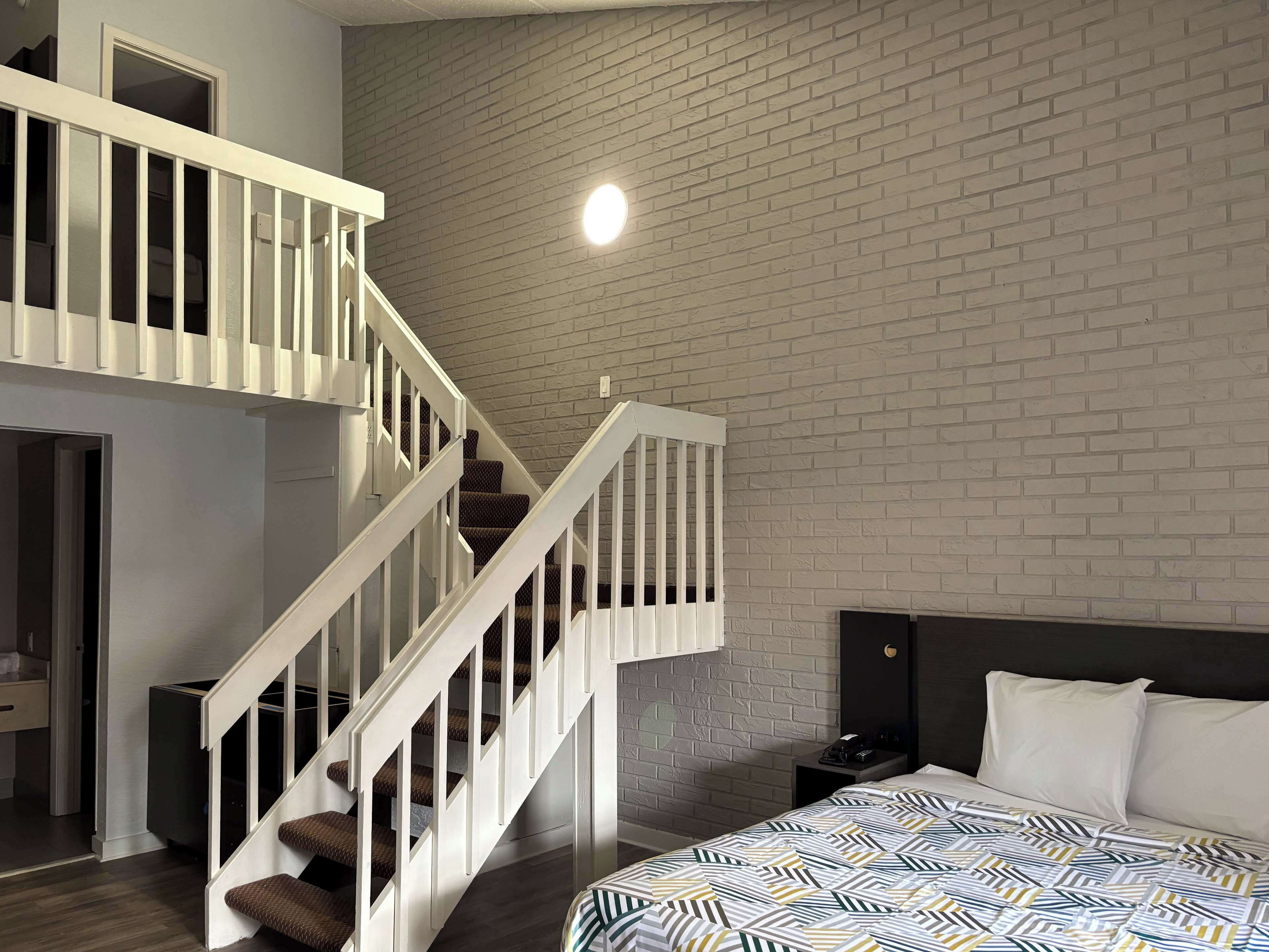 A modern bedroom with a brick-patterned wall, a white staircase, a bed with patterned bedding, and a nightstand with a lamp.