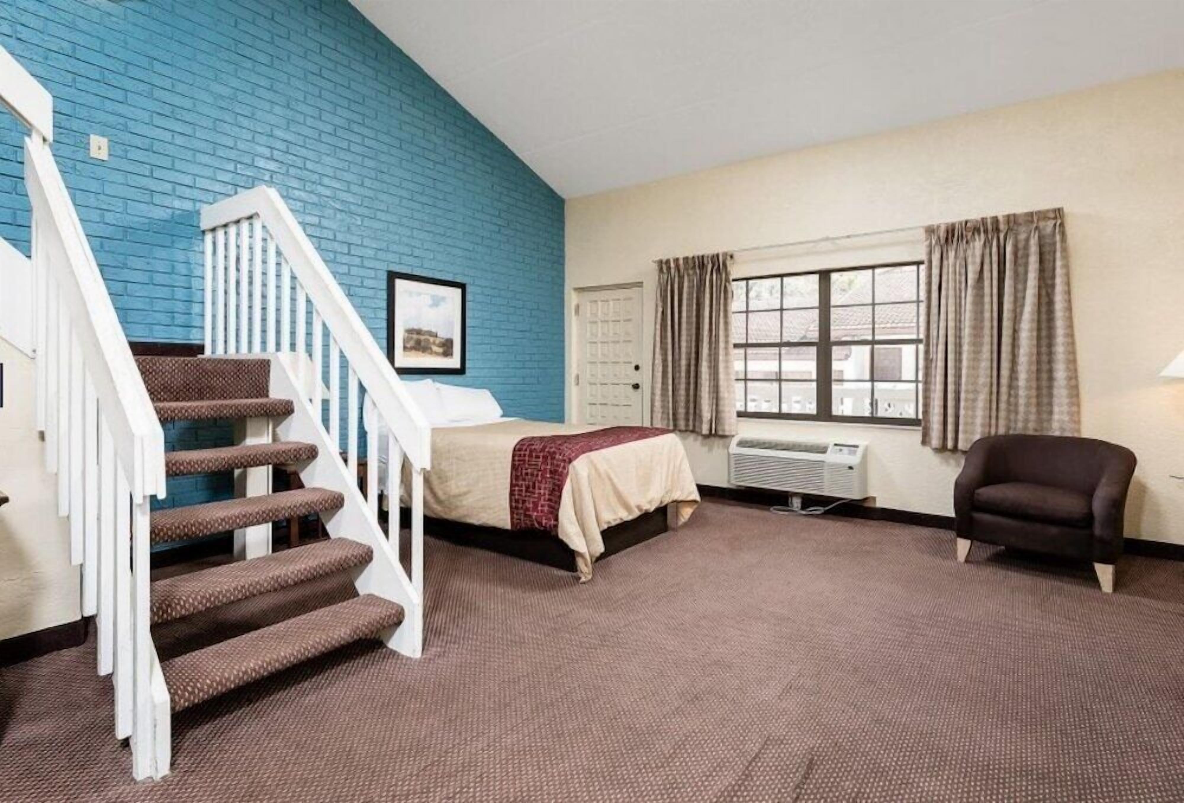 A hotel room with a blue brick wall, a bed with a red and white blanket, a brown armchair, a window with curtains, and a staircase with white railings.