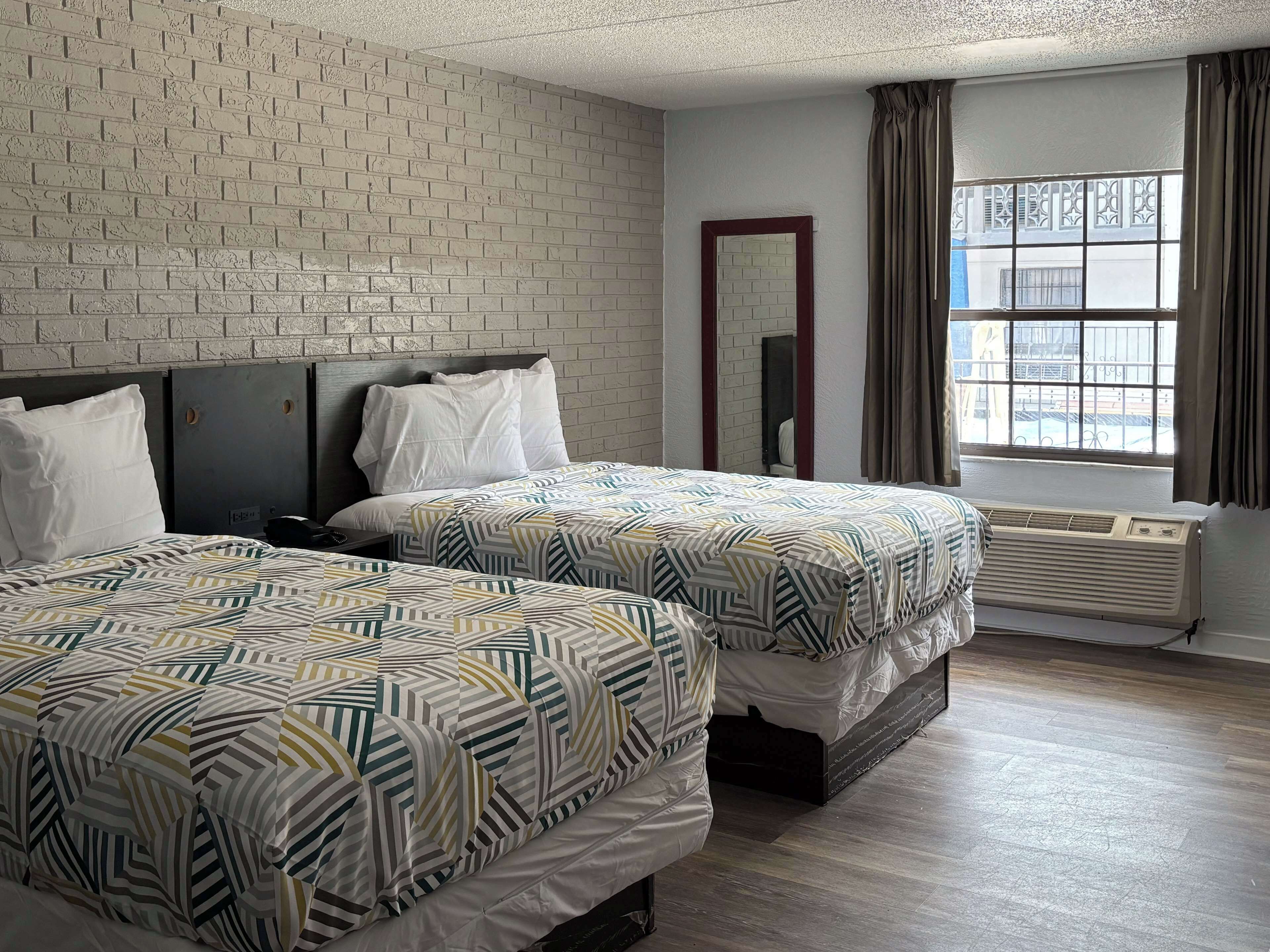 A hotel room with two beds, a brick wall, a window with curtains, and a mirror.