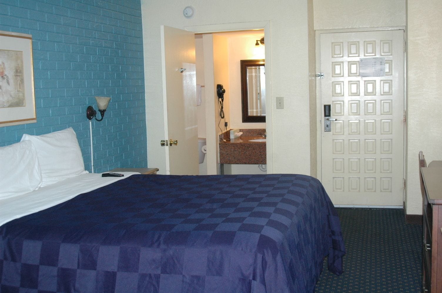 A hotel room with a bed, a desk, a chair, a bathroom with a mirror and sink, and a door with a lock.