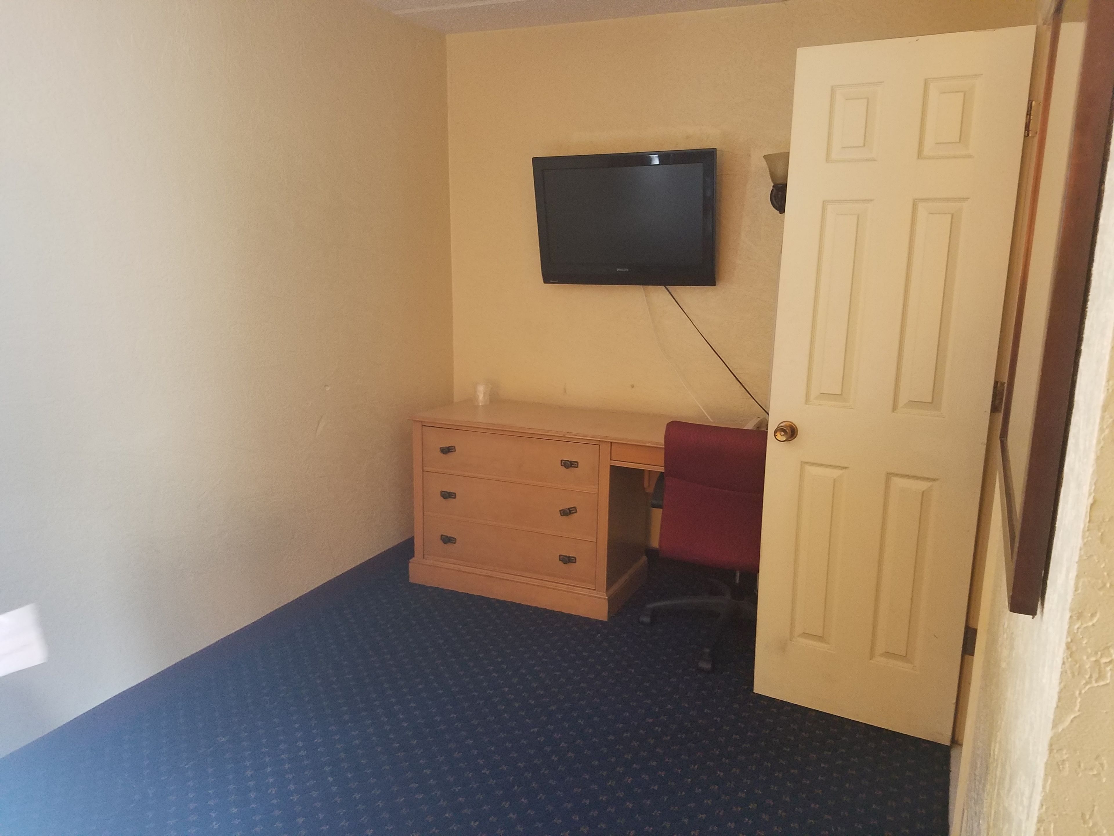 Una habitación pequeña con un escritorio de madera, una silla roja, un televisor montado en la pared y una puerta.