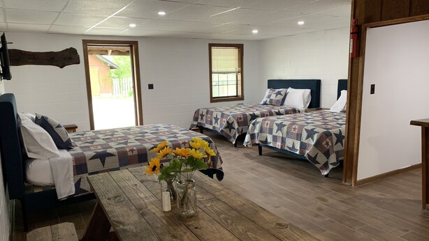 A hotel room with two beds, a wooden table with a vase of flowers, and a view of the outdoors through the windows. - Big South Fork Lodge (Jamestown)