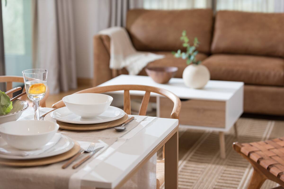 A dining area with a table set for two, a brown couch, and a small side table with a bowl and a vase.