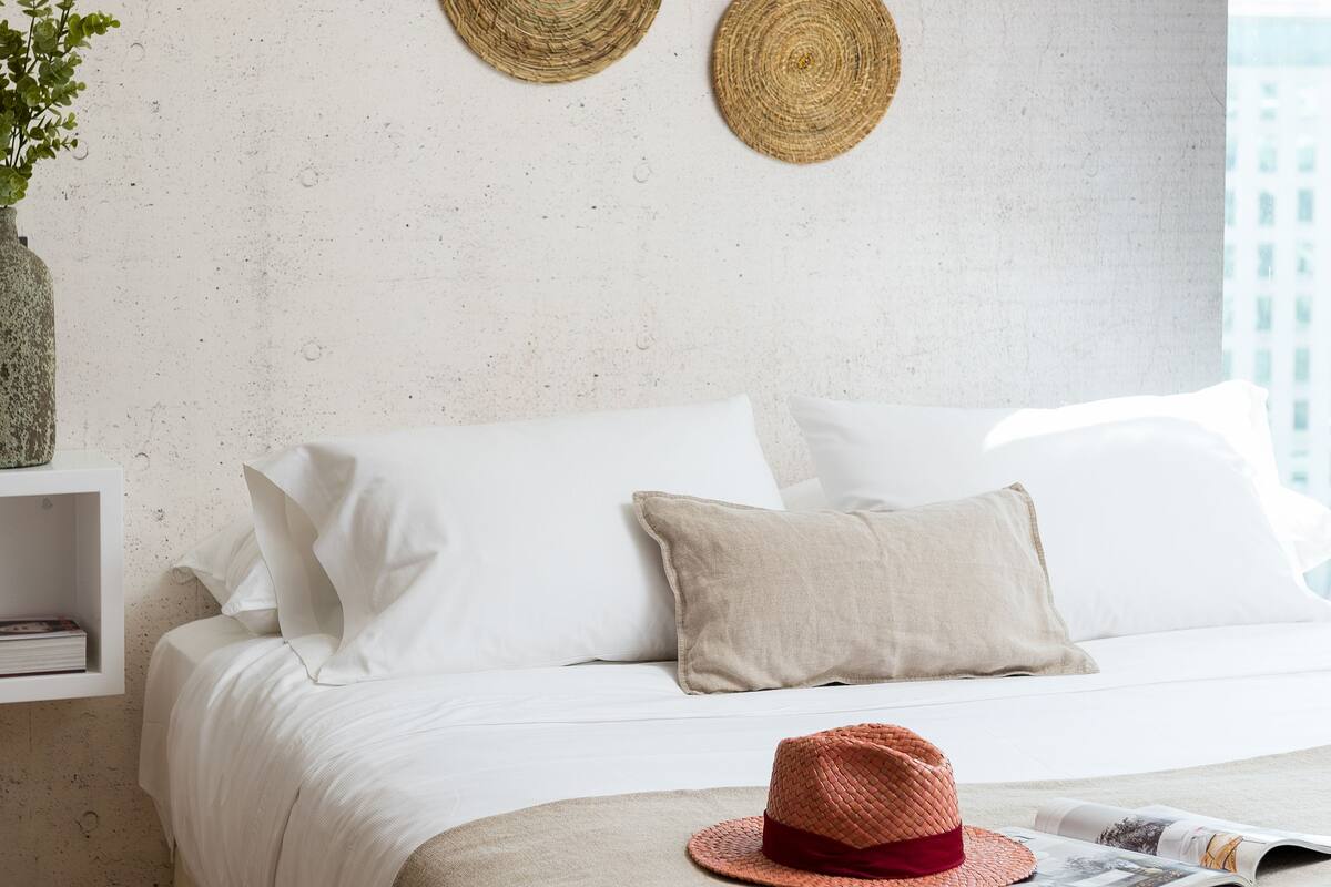 A neatly made bed with white bedding, a red woven hat, and a magazine on a beige throw.