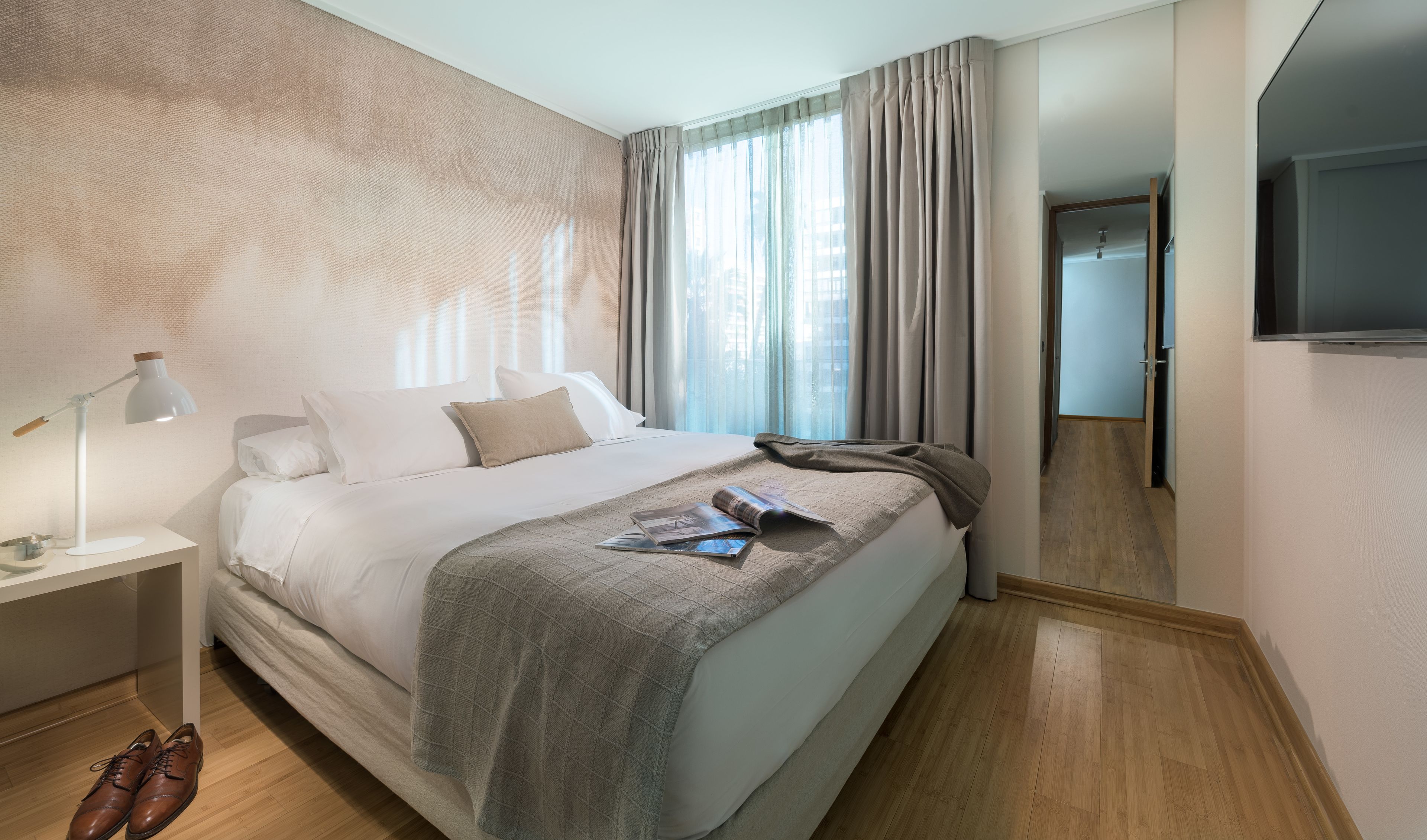 A neatly made bed with a magazine, a lamp, and a pair of shoes on the floor.